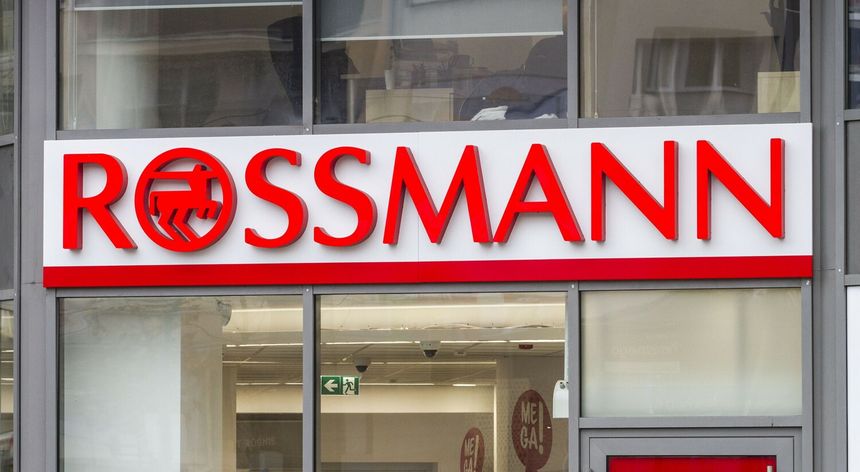 Znak firmowy Rossmann, czerwony napis na białym tle, z widocznymi witrynami sklepowymi.