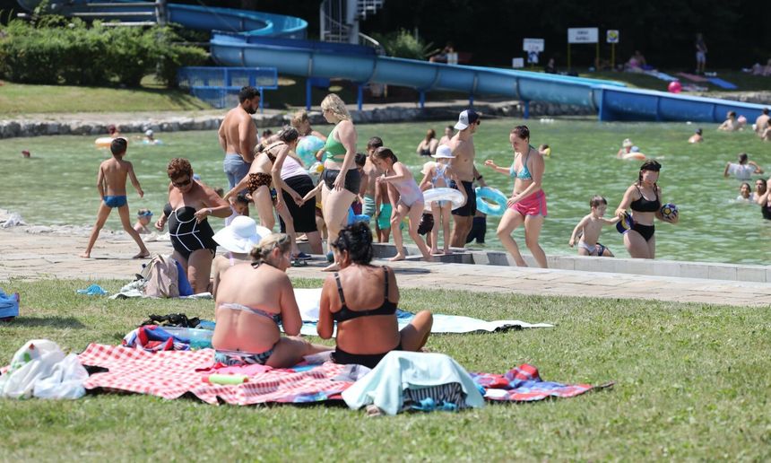 Lato nad wodą: ludzie bawią się w wodzie, leżą na trawie i rozkładają kocyki. Słońce.