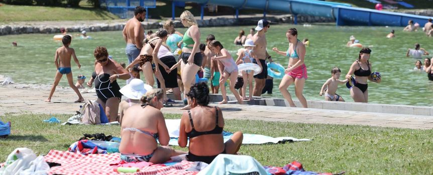 Lato nad wodą: ludzie bawią się w wodzie, leżą na trawie i rozkładają kocyki. Słońce.