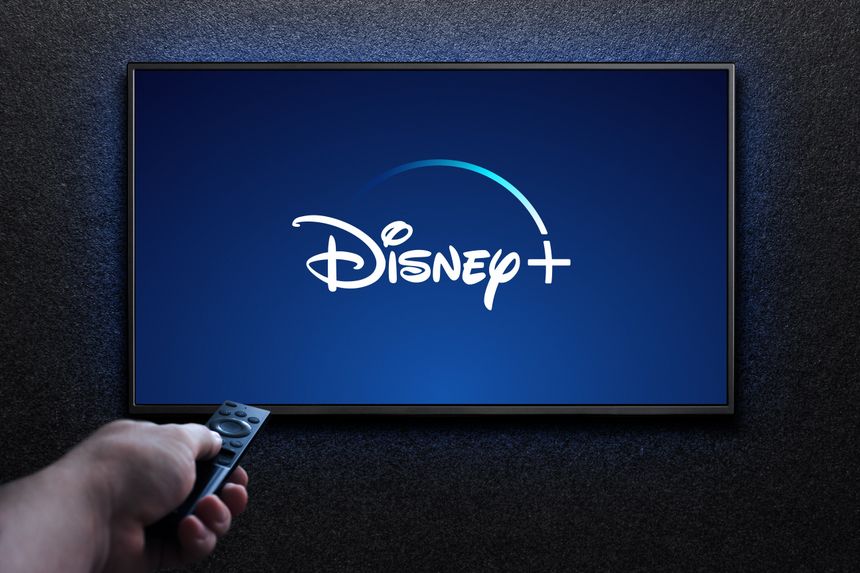 Na ekranie telewizora widoczny jest napis Disney+ na niebieskim tle. W ręku trzymany pilot.