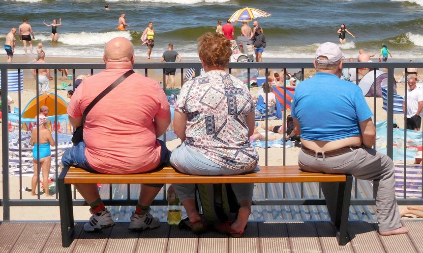Trzy osoby siedzą na ławce, patrząc na plażę i morze. Ludzie kąpią się i odpoczywają.