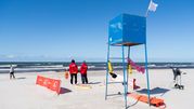 Na plaży z ratownikami w czerwonych strojach, wieżą ratunkową i morzem w tle.