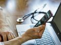 Osoba trzyma telefon, korzystając z laptopa, na stole leży stetoskop.