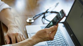 Osoba trzyma telefon, korzystając z laptopa, na stole leży stetoskop.