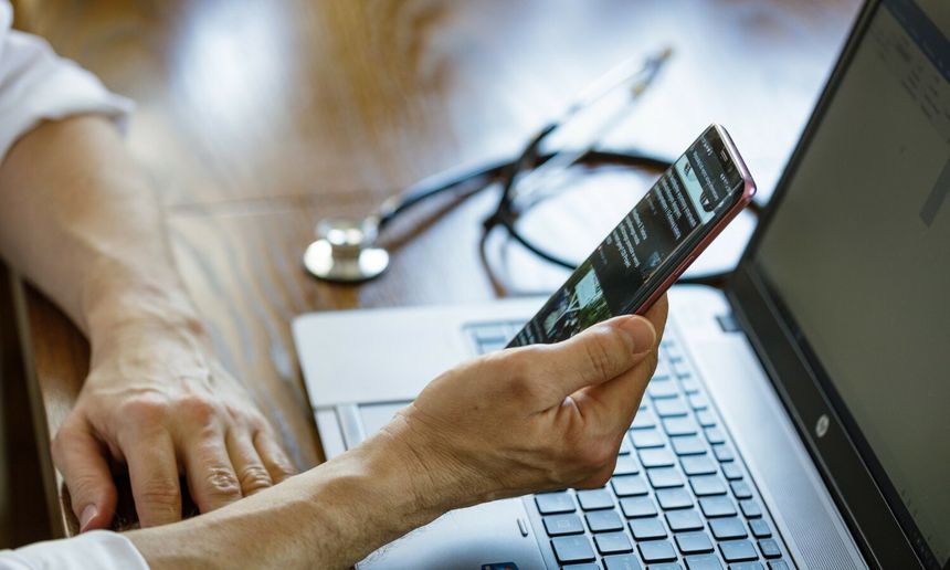 Osoba trzyma telefon, korzystając z laptopa, na stole leży stetoskop.
