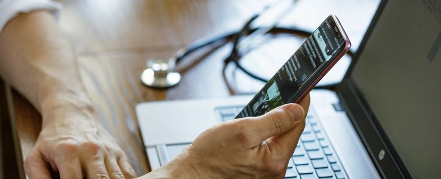 Osoba trzyma telefon, korzystając z laptopa, na stole leży stetoskop.