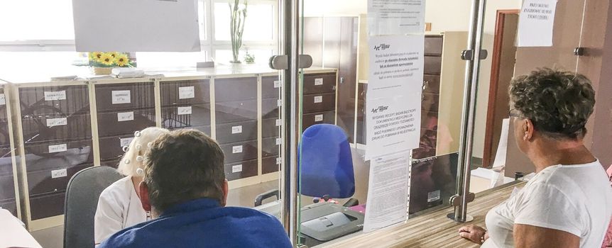 Na zdjęciu ludzie czekający przy oknie, z tablicą informującą o ograniczeniach.