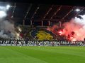 Kibice na stadionie Pogoni Szczecin, w tle ogromna flaga, dym i transparent.
