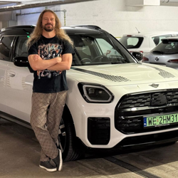 Mężczyzna z długimi włosami stoi przy białym samochodzie w parkingu.