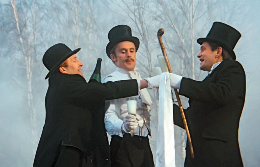 Trzech mężczyzn w kapeluszach toczą kieliszki w dymnej scenerii leśnej.