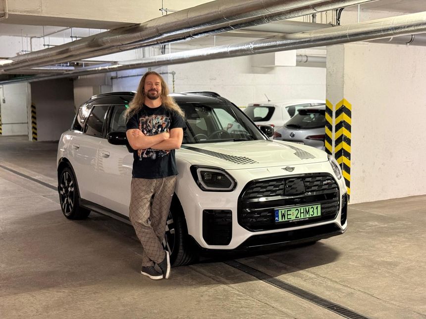 Mężczyzna z długimi włosami stoi obok białego samochodu w parkingu.