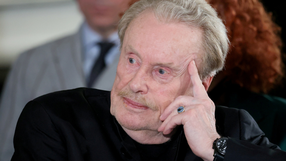 Mężczyzna z siwymi włosami i wąsem, w czarnym ubraniu, myśli, opierając głowę na dłoni.
