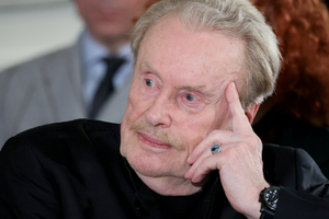 Mężczyzna z siwymi włosami i wąsem, w czarnym ubraniu, myśli, opierając głowę na dłoni.