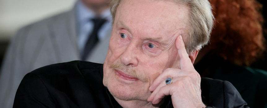 Mężczyzna z siwymi włosami i wąsem, w czarnym ubraniu, myśli, opierając głowę na dłoni.