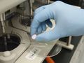 Ręka w niebieskiej rękawicy trzyma fioletową tabletkę nad laboratoryjnym urządzeniem.