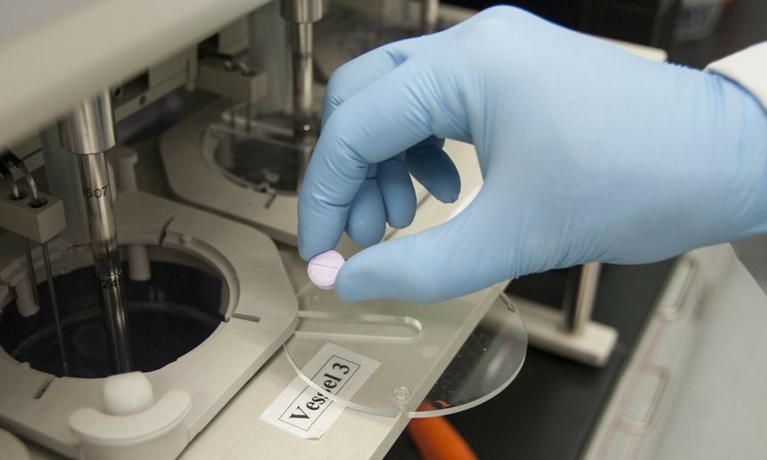 Ręka w niebieskiej rękawicy trzyma fioletową tabletkę nad laboratoryjnym urządzeniem.