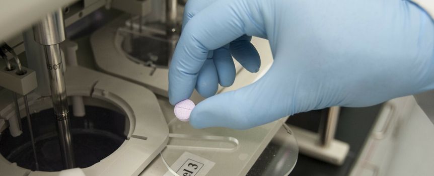 Ręka w niebieskiej rękawicy trzyma fioletową tabletkę nad laboratoryjnym urządzeniem.