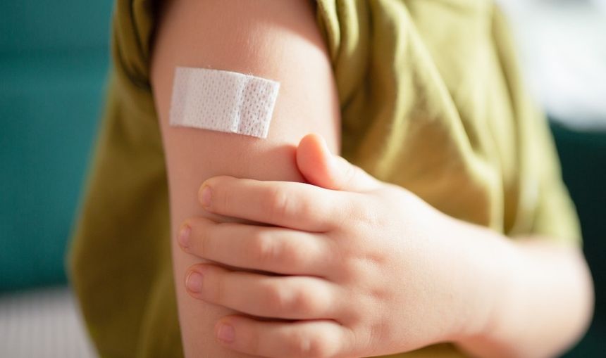Dziecięca ręka z opatrunkiem na przedramieniu, trzymana drugą ręką. Tło rozmyte.
