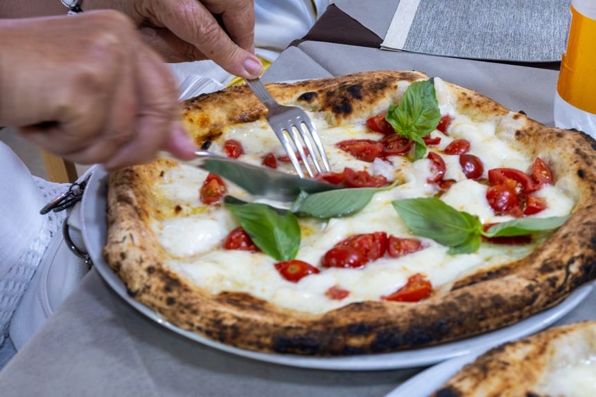 Na talerzu leży pizza z serem, pomidorami i bazylią. Ktoś kroi ją widelcem i nożem.