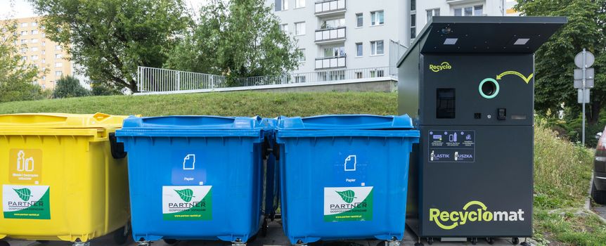 Kolorowe kontenery na odpady i automat do recyclingu w tle, w miejskim otoczeniu.