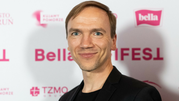 Mężczyzna w czarnej marynarce uśmiecha się na tle kolorowych napisów na ściance.