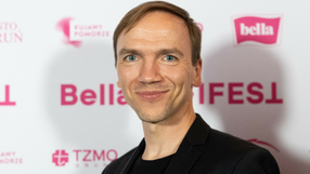 Mężczyzna w czarnej marynarce uśmiecha się na tle kolorowych napisów na ściance.
