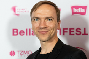 Mężczyzna w czarnej marynarce uśmiecha się na tle kolorowych napisów na ściance.