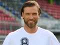 Mężczyzna z brodą w białym T-shircie, uśmiecha się na tle pustych trybun stadionu.