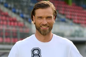 Mężczyzna z brodą w białym T-shircie, uśmiecha się na tle pustych trybun stadionu.
