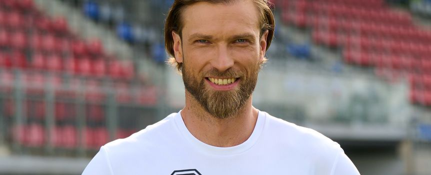 Mężczyzna z brodą w białym T-shircie, uśmiecha się na tle pustych trybun stadionu.