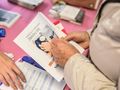 Osoba trzyma ulotkę o zdrowiu, na stole widoczne materiały informacyjne. Tło różowe.