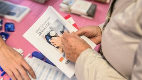 Osoba trzyma ulotkę o zdrowiu, na stole widoczne materiały informacyjne. Tło różowe.