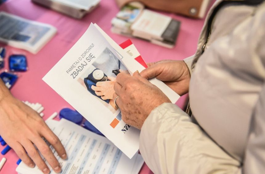 Osoba trzyma ulotkę o zdrowiu, na stole widoczne materiały informacyjne. Tło różowe.