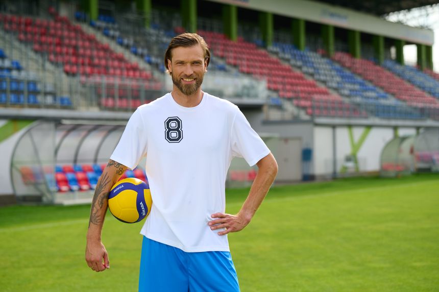 Mężczyzna w białej koszulce z numerem 8, trzyma piłkę, stoi na boisku. W tle trybuny.