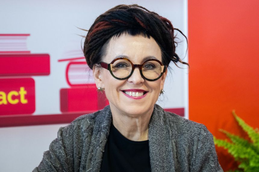 Kobieta z okularami, w szaro-czarnym swetrze, uśmiecha się w kolorowym otoczeniu.