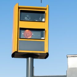 Drogowy fotoradar w żółtej obudowie na tle nieba. Czerwone światło włączone.