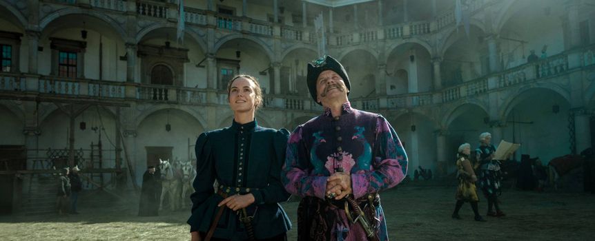 Dwoje ludzi w strojach historycznych, stojących w dziedzińcu budynku, z uśmiechami na twarzach.