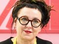 Olga Tokarczuk. Kobieta z okularami, czarnymi włosami w dredach, uśmiecha się na różowym tle.