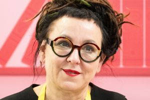 Olga Tokarczuk. Kobieta z okularami, czarnymi włosami w dredach, uśmiecha się na różowym tle.