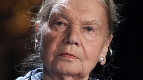 Na zdjęciu znajduje się poważnie wyglądająca starsza kobieta z jasnymi włosami i kolczykami.