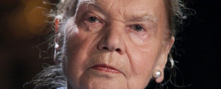 Na zdjęciu znajduje się poważnie wyglądająca starsza kobieta z jasnymi włosami i kolczykami.