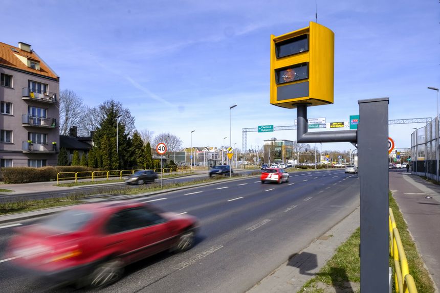 Na zdjęciu droga z fotoradarem. W tle budynki i znaki drogowe. Ruchome samochody.
