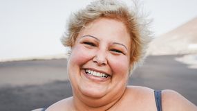 Uśmiechnięta kobieta z blond włosami stoi na tle plaży, w promieniach słońca.