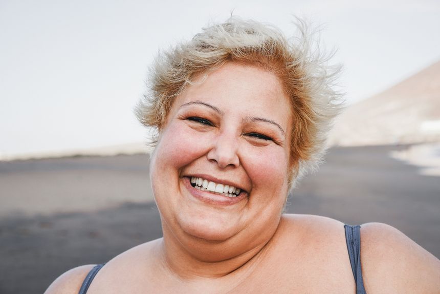Uśmiechnięta kobieta z blond włosami stoi na tle plaży, w promieniach słońca.