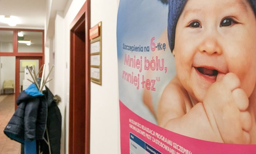 Plakat w korytarzu z uśmiechniętym niemowlęciem i hasłem o szczepieniach. W tle wieszaki z odzieżą.