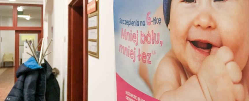 Plakat w korytarzu z uśmiechniętym niemowlęciem i hasłem o szczepieniach. W tle wieszaki z odzieżą.