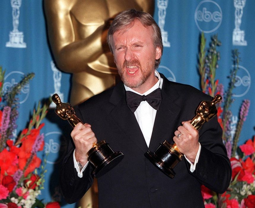 James Cameron z 2 Oscarami. Mężczyzna w garniturze trzyma dwa złote oscarowe statuetki, wyrażając radość. Tło kwiatowe.