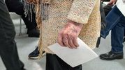 Na zdjęciu widoczna jest długa ręka trzymająca kartkę, w tle inni ludzie siedzący na krzesłach.