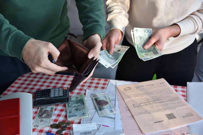 Dwie osoby wymieniają pieniądze przy stole, obok kalkulator, dokumenty i monety.