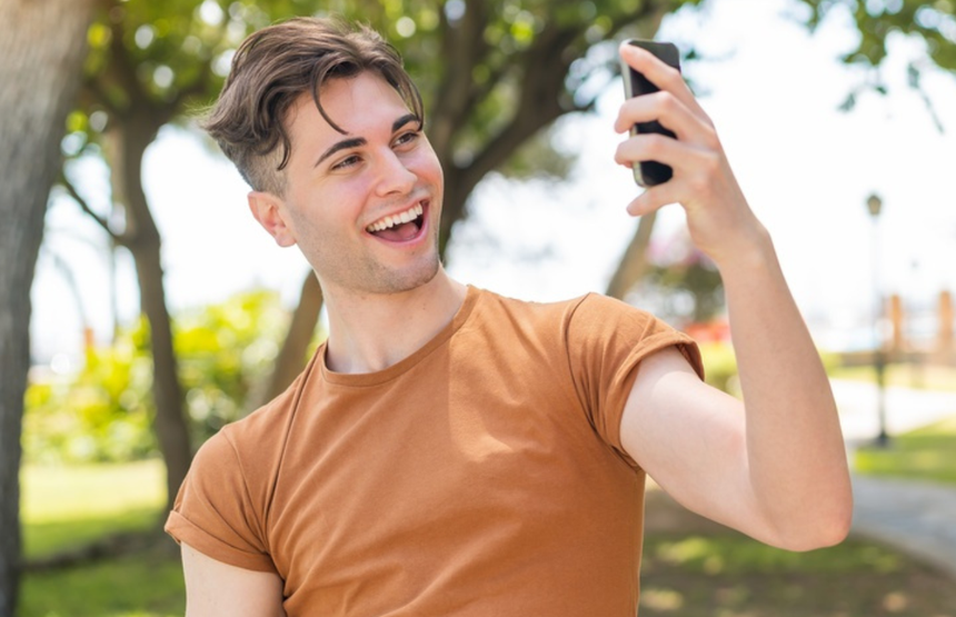 Młody mężczyzna uśmiecha się i robi selfie w parku, otoczony zielenią.